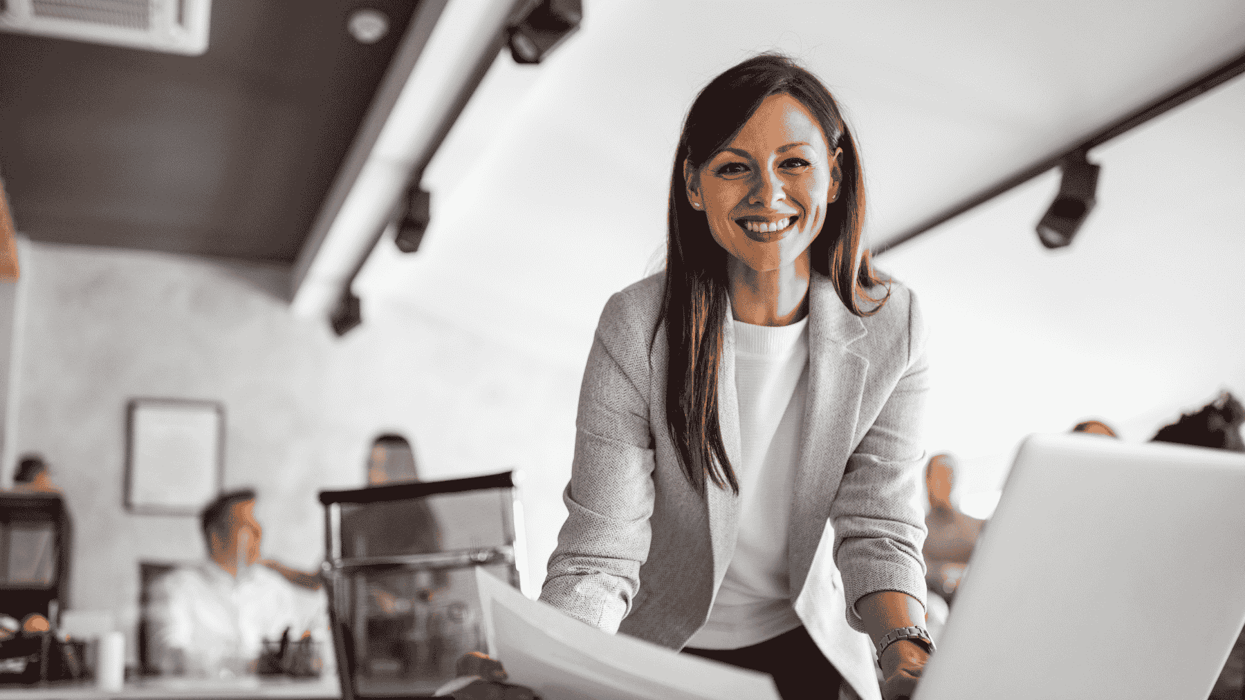 Femeie zambind la camera cu laptop in fata si hartii in mana